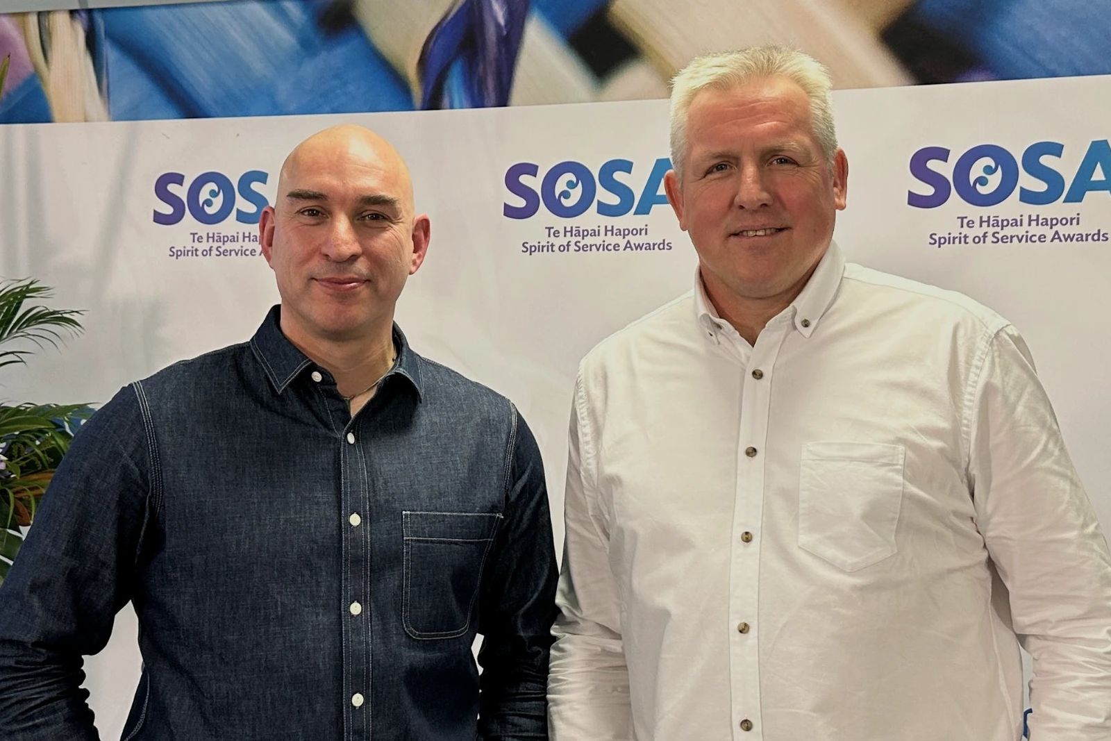 Two men in casual shirts stand side by side smiling slightly in front of an awards backdrop, with a flower arrangement at left.
