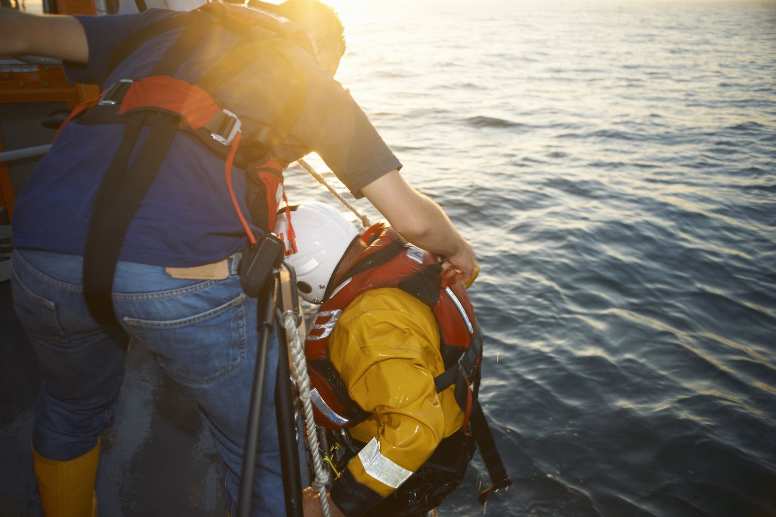 A man on a boat deck leans over the side to help a rescuer in a yellow jacket, life vest and white helmet at the sea surface.