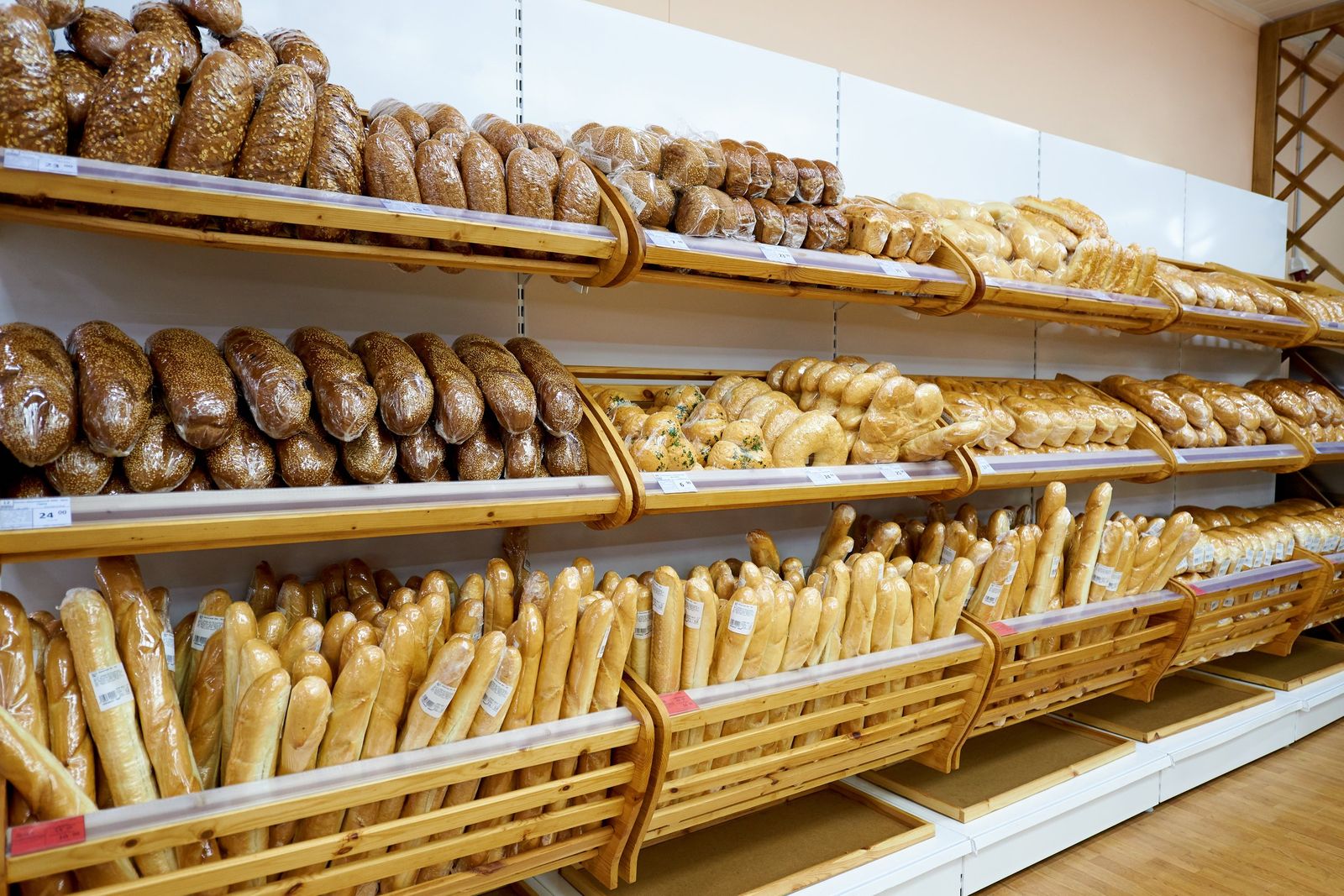 Wooden shelves in a shop packed with wrapped loaves, seeded rolls and long baguettes, with price tags along the shelf edges.