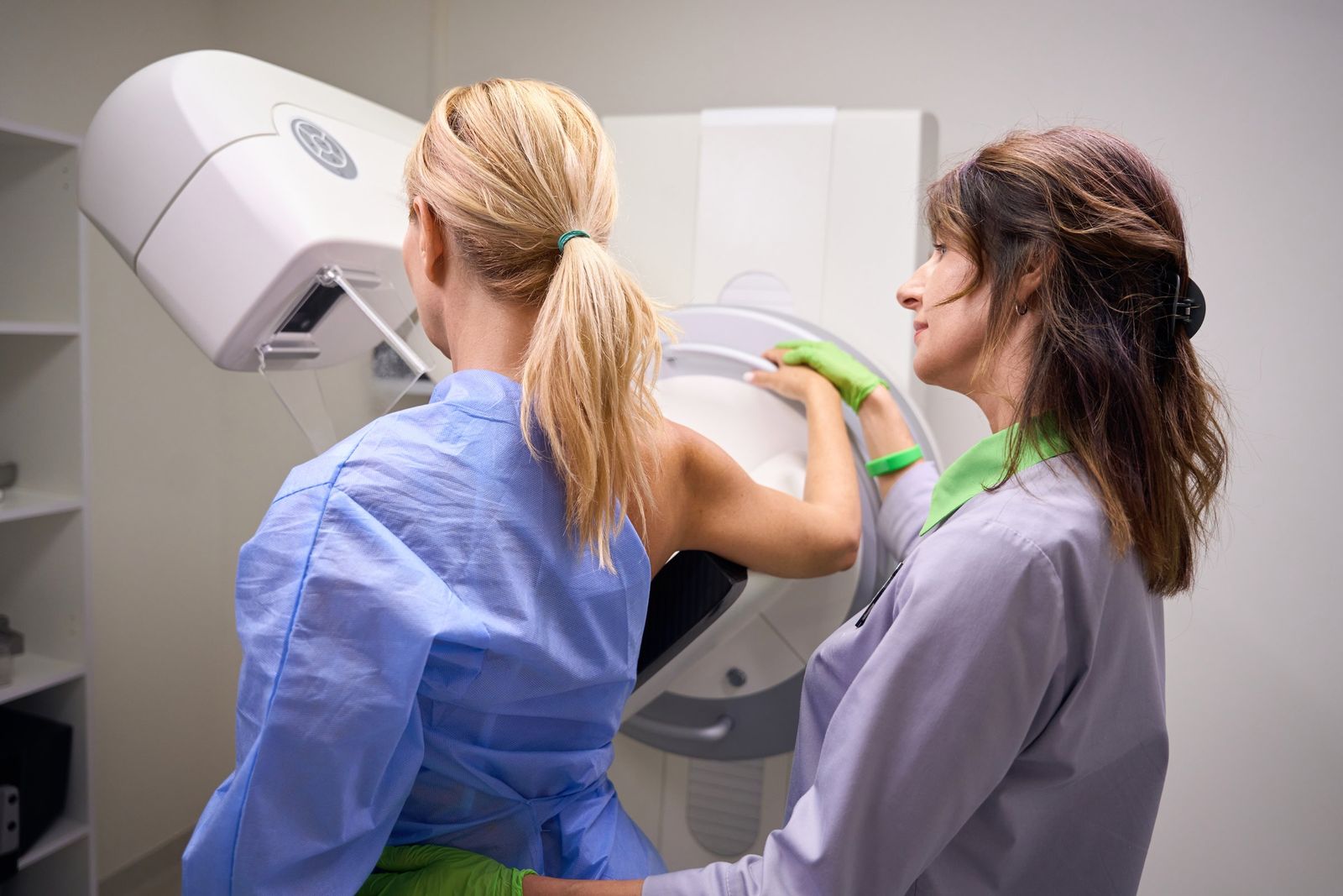A woman in a blue gown stands at a mammogram machine while a radiographer in gloves guides her into position.