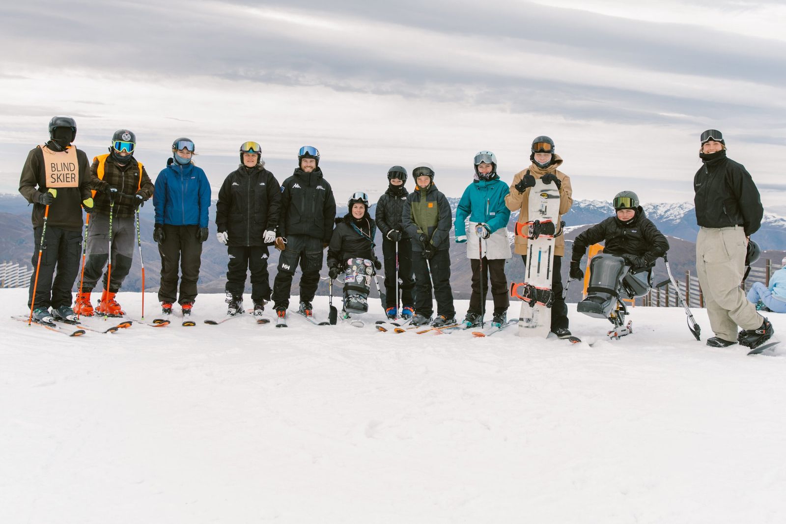 A row of twelve skiers, a snowboarder and a sit-ski user in winter gear pose together on a snowy mountain slope.