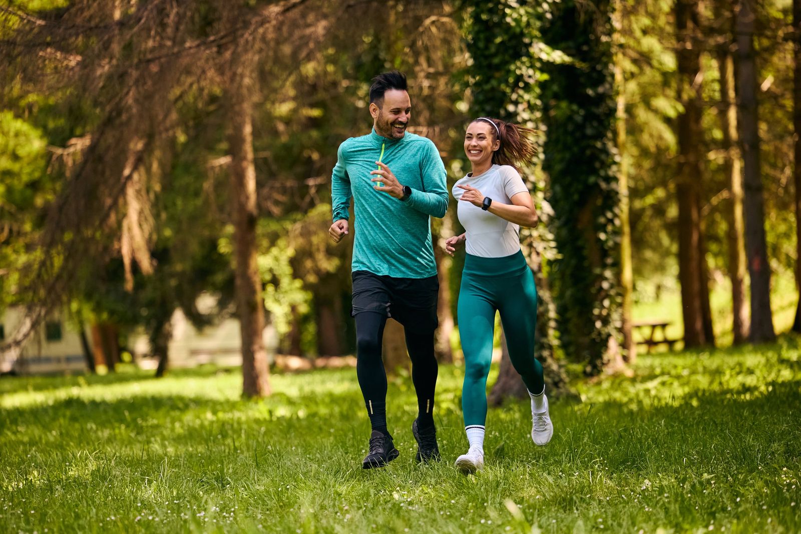 A smiling man and woman in activewear jog side by side across a grassy clearing in a sunlit pine forest.