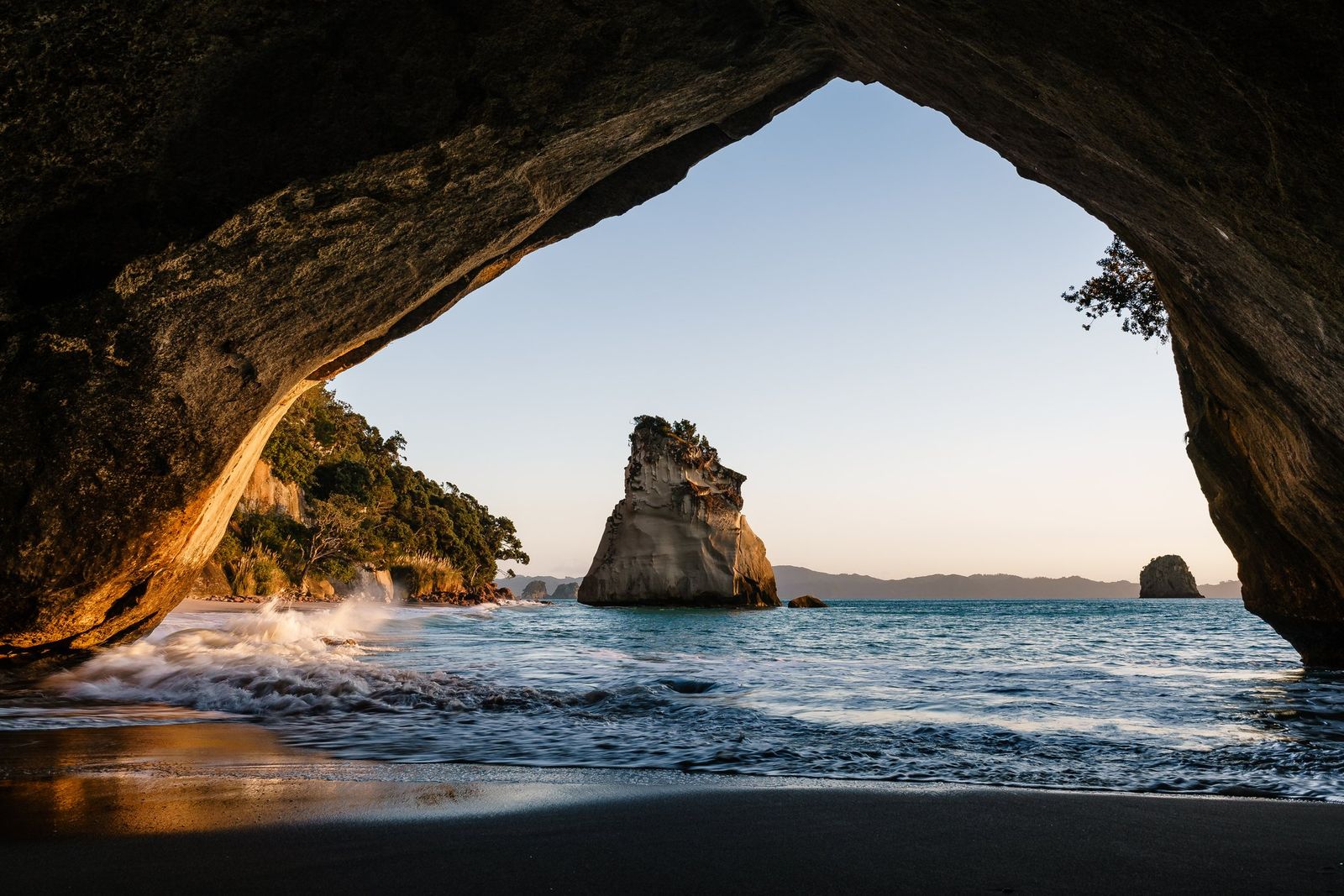 A large rock stack stands in calm sea, framed by a dark sea cave arch, with waves washing onto a dark sandy beach.
