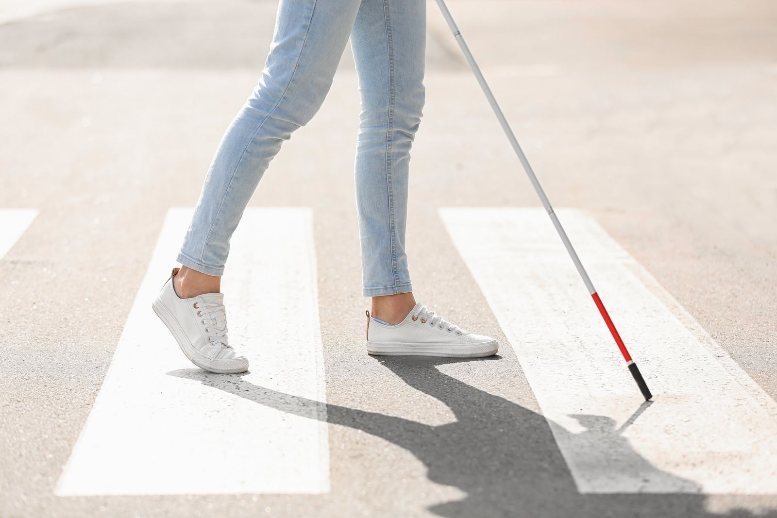 Person in light jeans and white sneakers walks across a pedestrian crossing, tapping a red and white cane on the road.