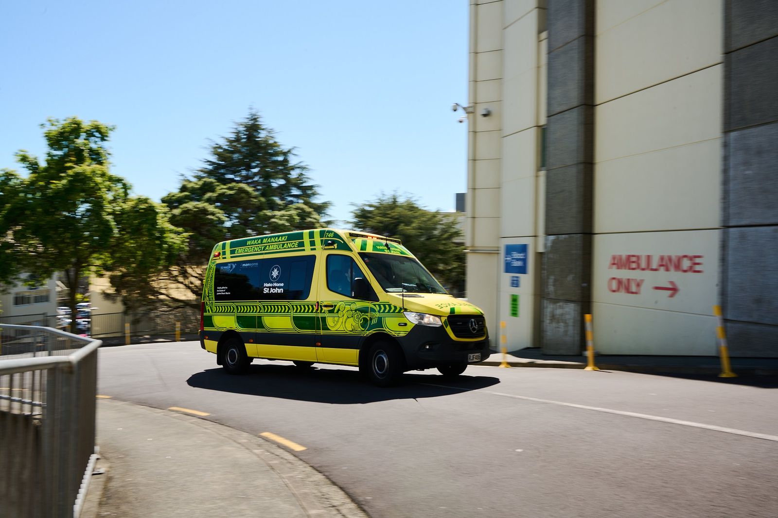 A yellow and green ambulance drives round a curve beside a hospital building marked ambulance only, on a sunny day.