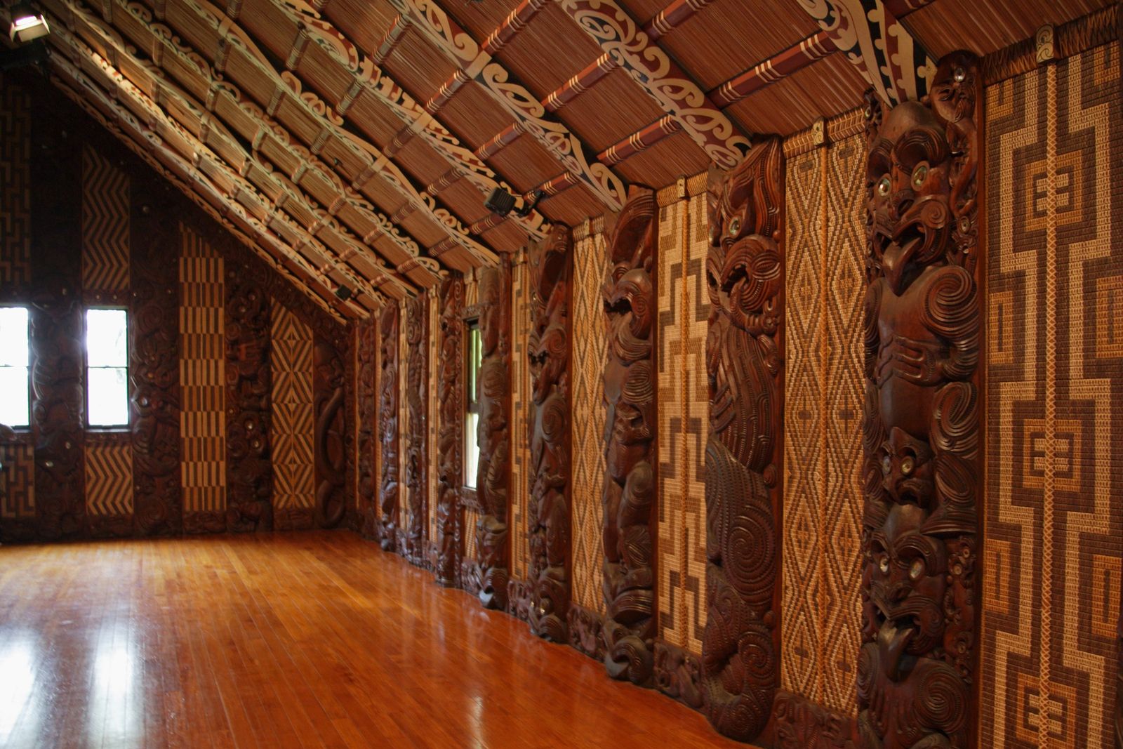 Empty meeting house interior with polished wooden floor, carved wooden figures with shell eyes, woven wall panels and patterned rafters.