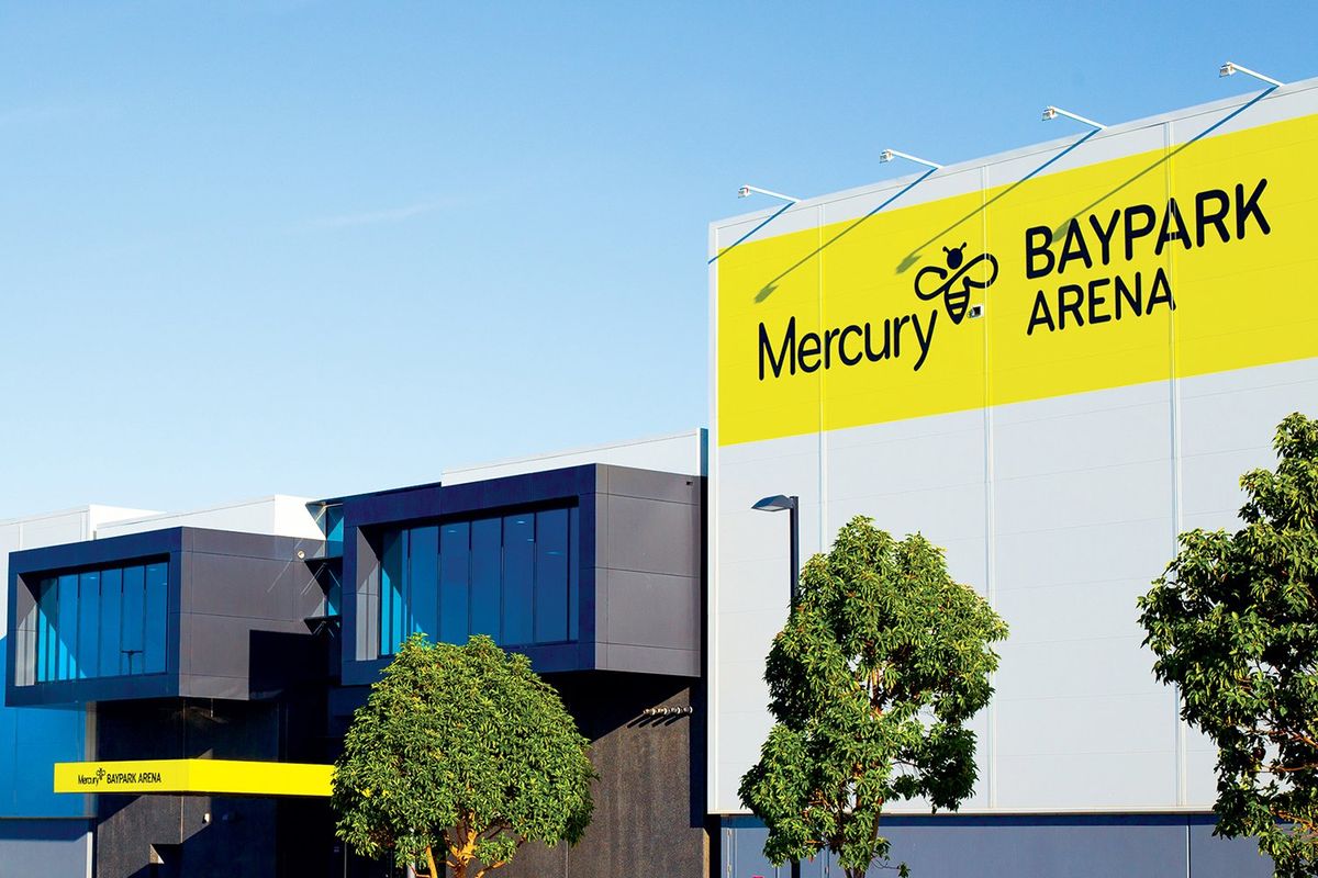 Exterior of a modern arena building with a large yellow sign featuring a bee logo, dark glass sections and green trees under a clear blue sky.