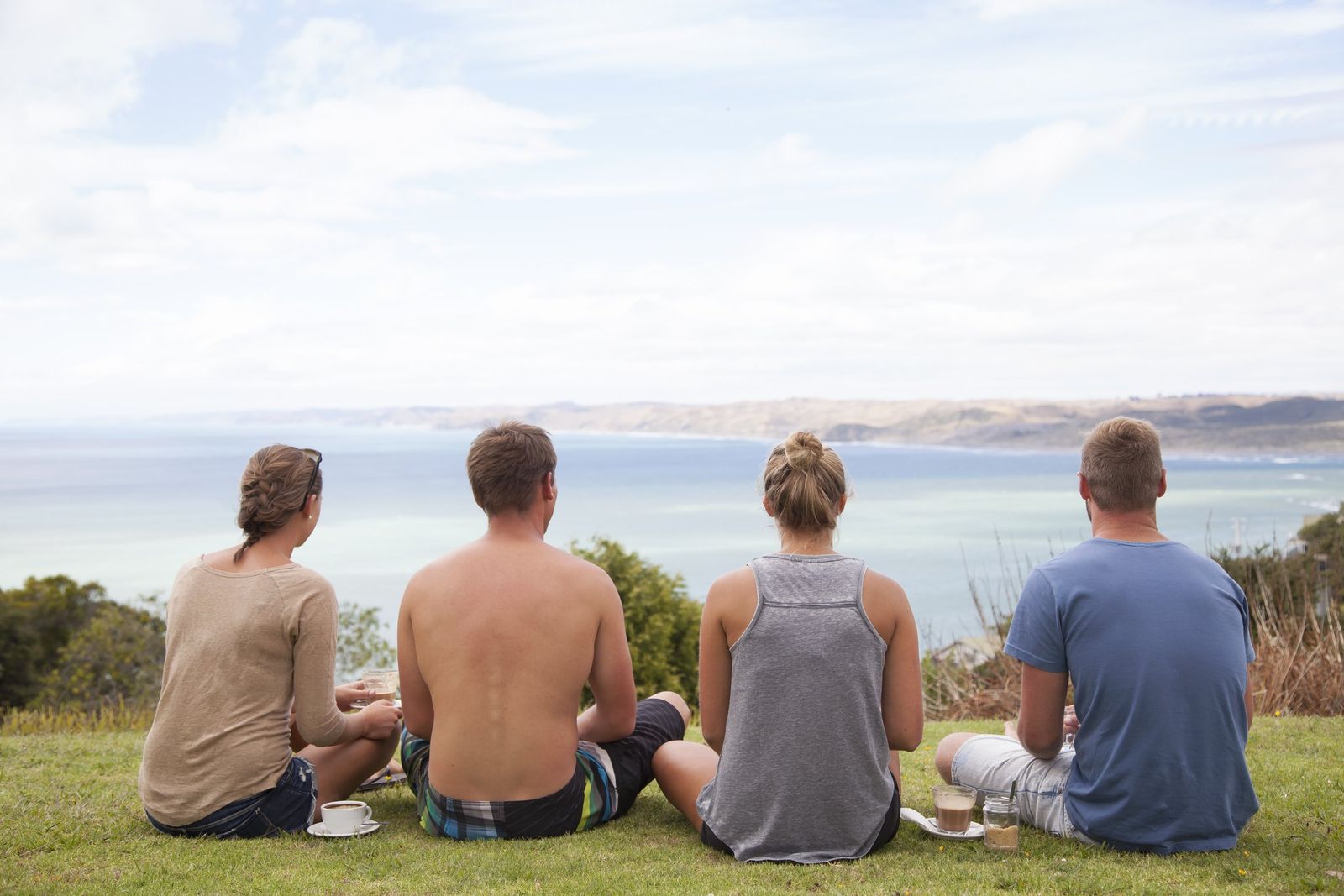 Four friends sitting on the grass above the sea with coffee, looking out at the view.
