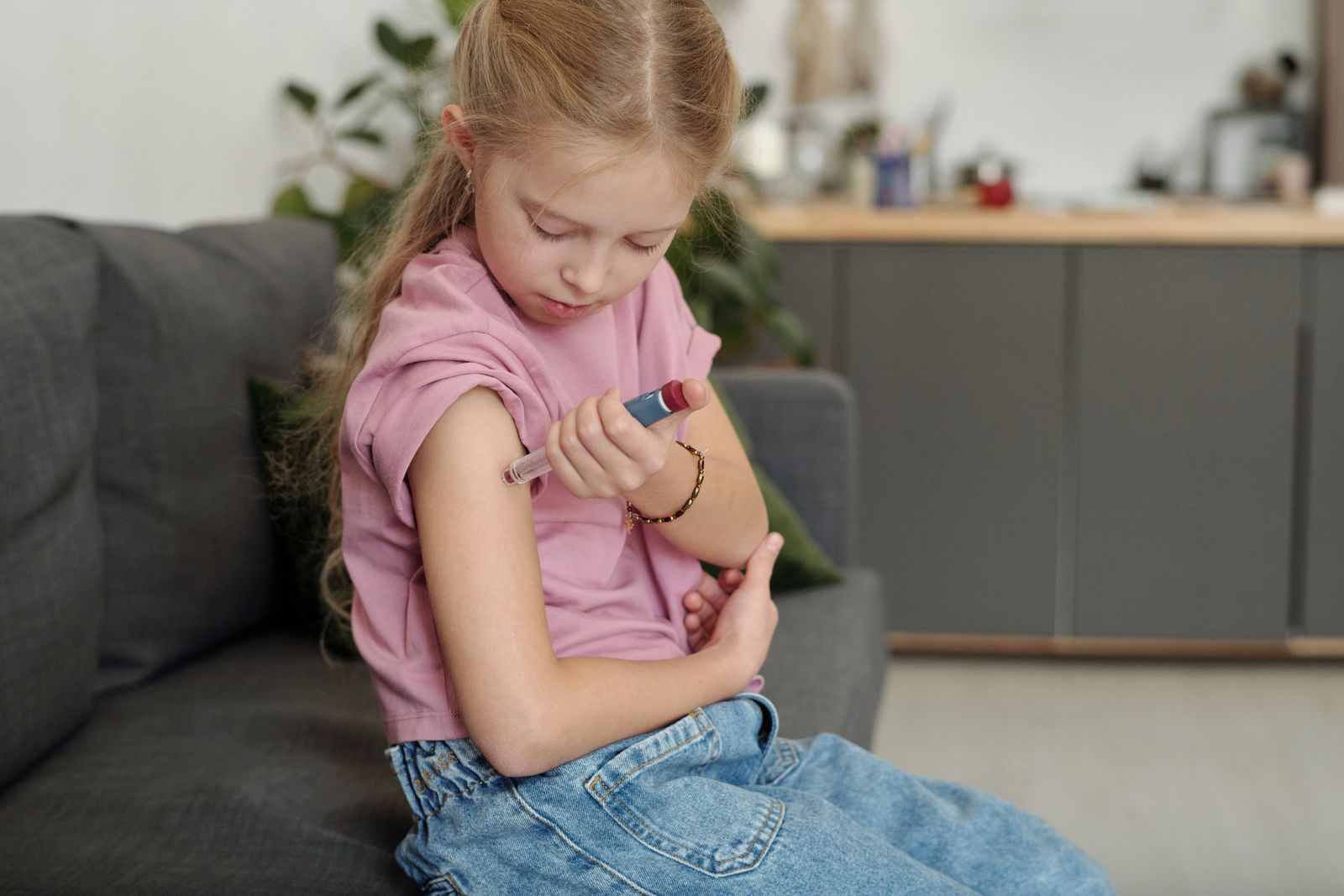 A young girl with long fair hair sits on a grey sofa at home, holding an insulin pen against her upper arm.