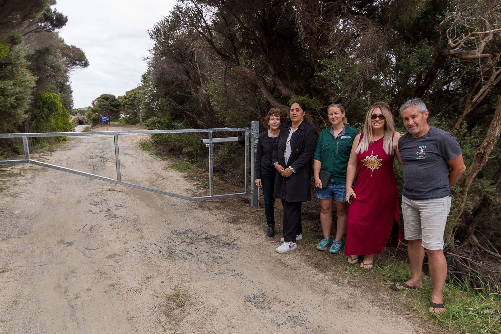 Five people stand together beside a new metal boom gate across a sandy track lined with scrubby native bush.