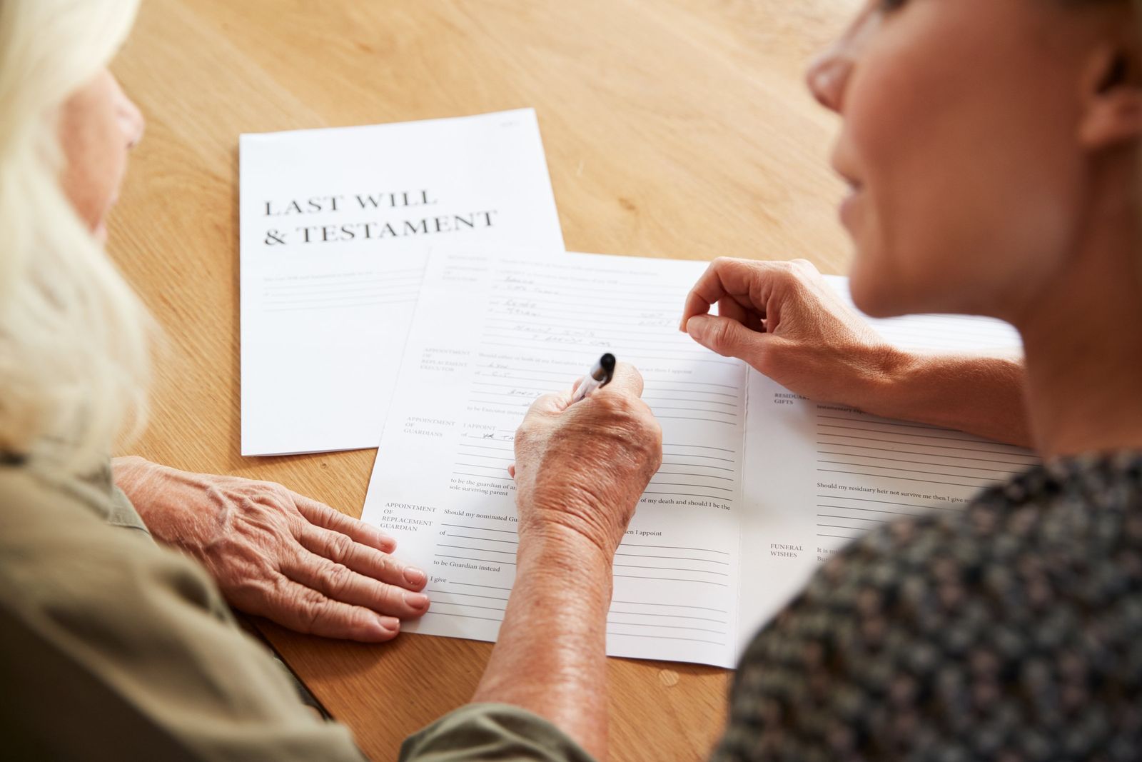 Two women sit at a wooden table as one with white hair signs a will, a Last Will and Testament cover beside it.