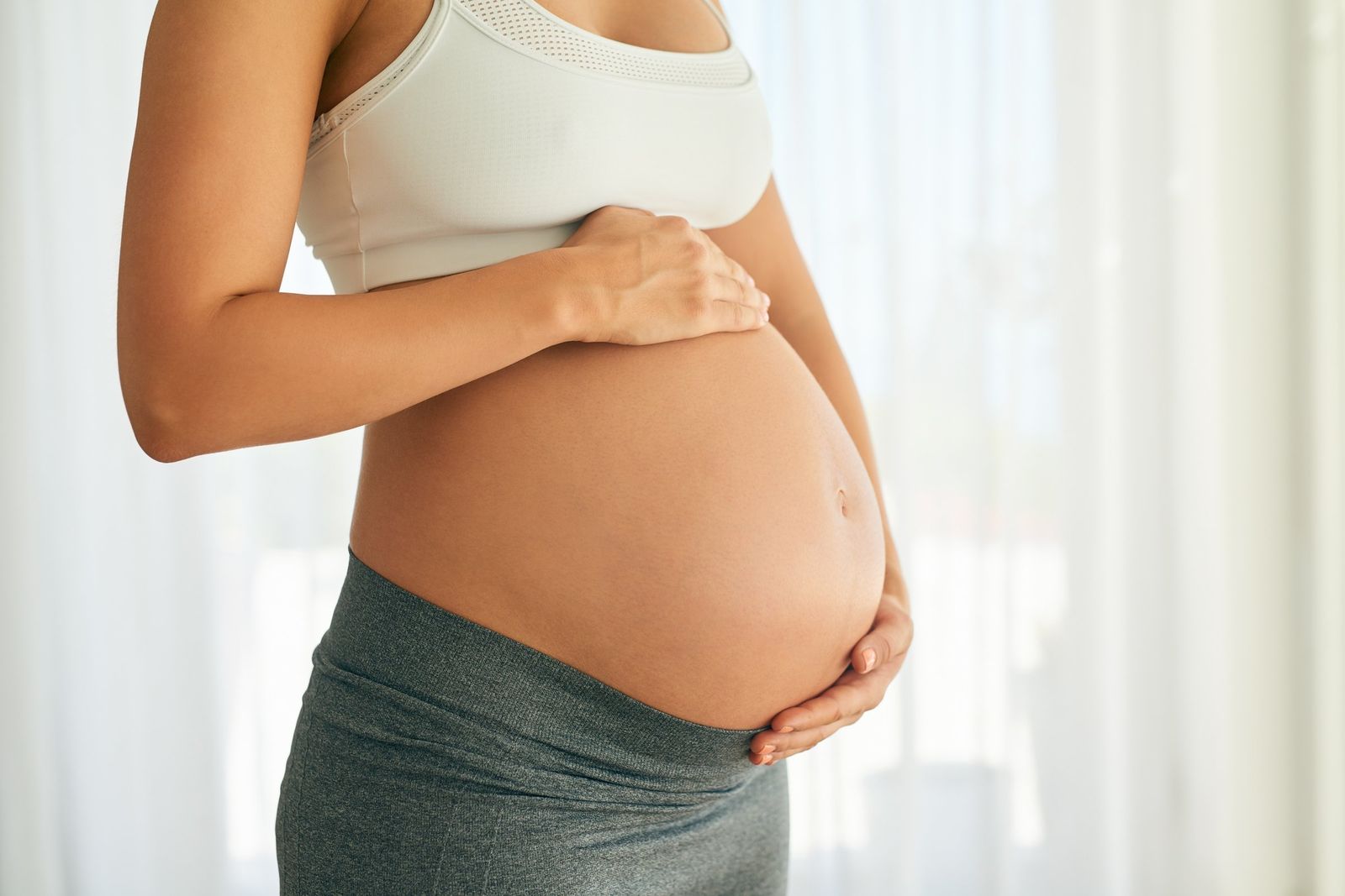 A pregnant woman in a white sports top and grey leggings cradles her bare belly with both hands beside a bright curtained window.