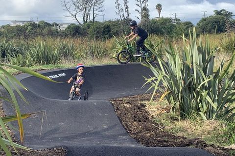 A young child on a balance bike and an older rider on a bicycle ride around a sealed pump track surrounded by flax.