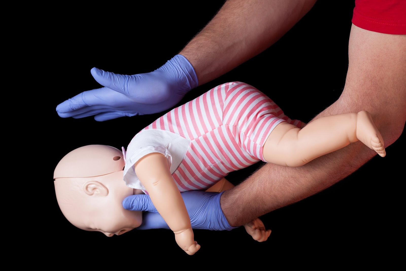 Gloved hands hold a baby training doll face down along a forearm, with the other hand ready to give back blows.