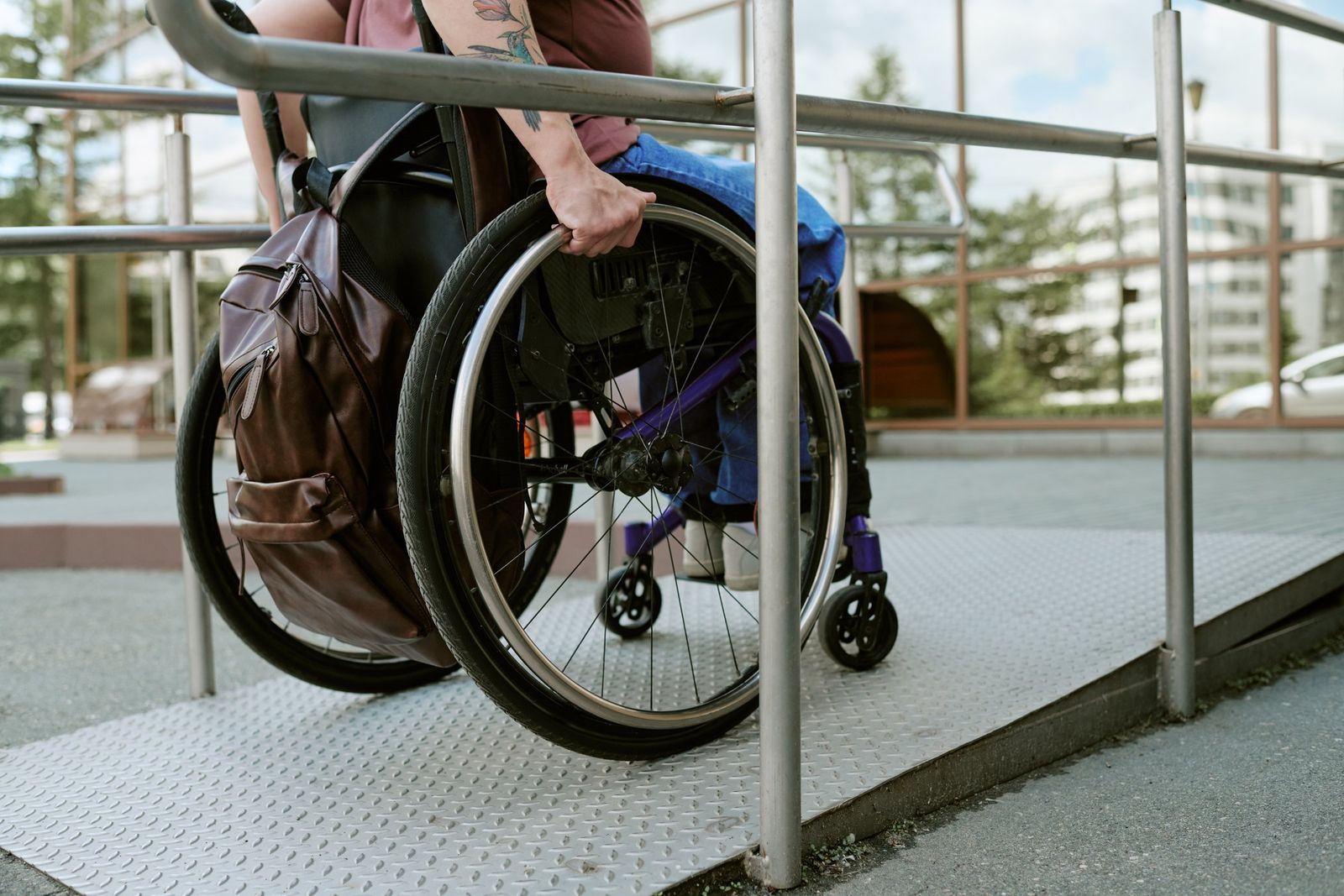 A person with a tattooed arm pushes a wheelchair with a brown backpack up a metal ramp with handrails outside a building.