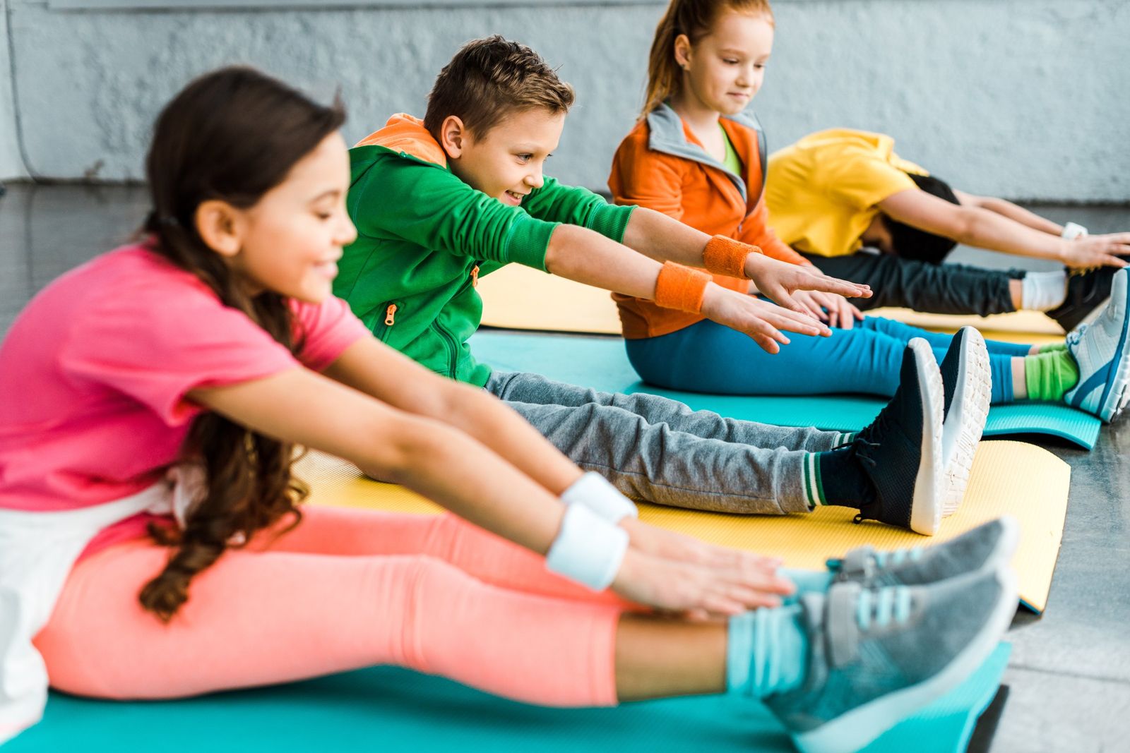 Four smiling children in bright sportswear sit on colourful gym mats, stretching their arms forward towards their toes in a row.