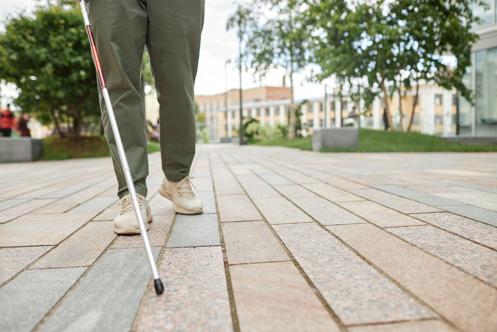 Person in olive trousers and cream sneakers walks along a paved path in a park, sweeping a white cane ahead.